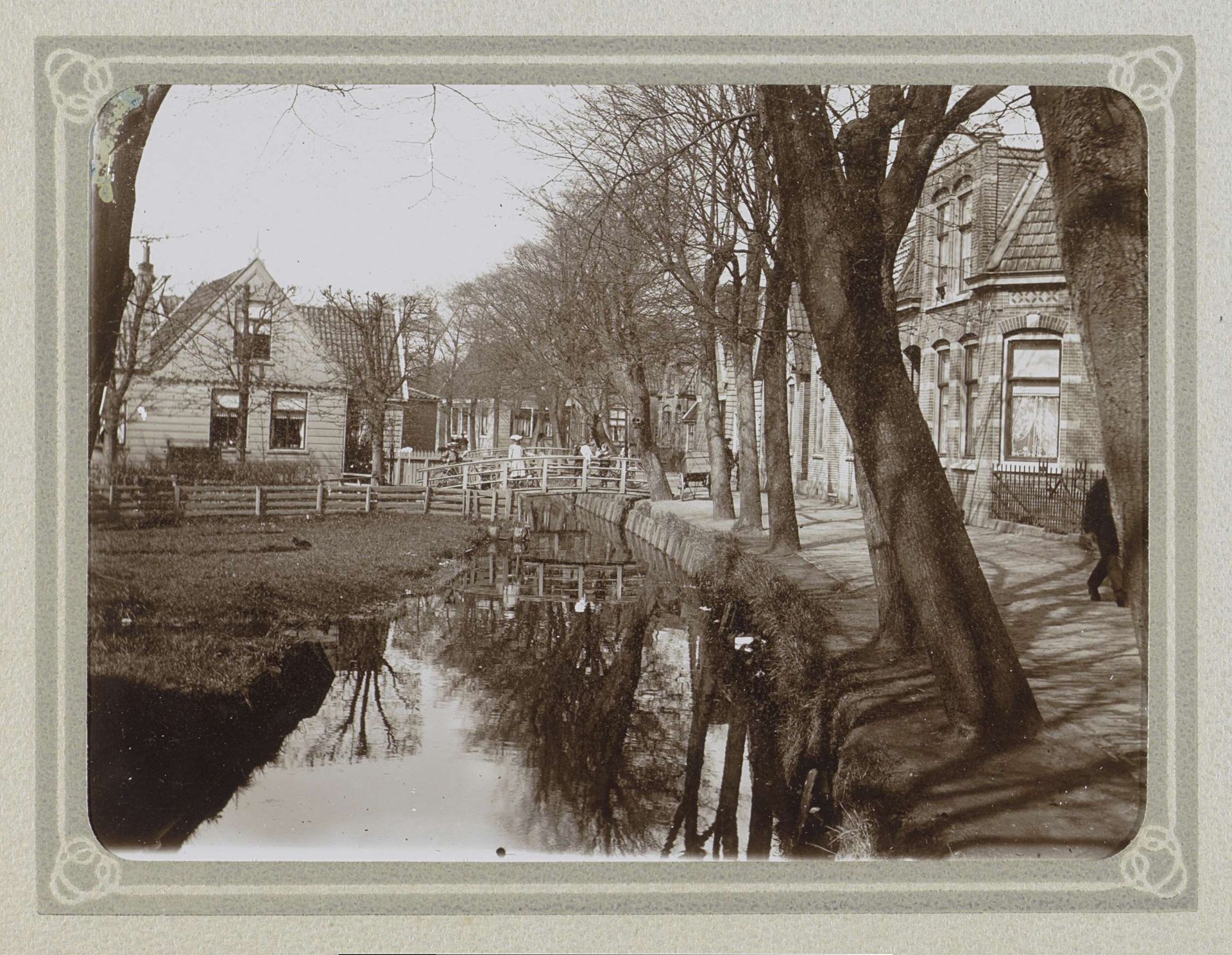 Dorpsstraat met huizen, een sloot en kinderen met fietsen op een brug by Folkert Idzes de Jong