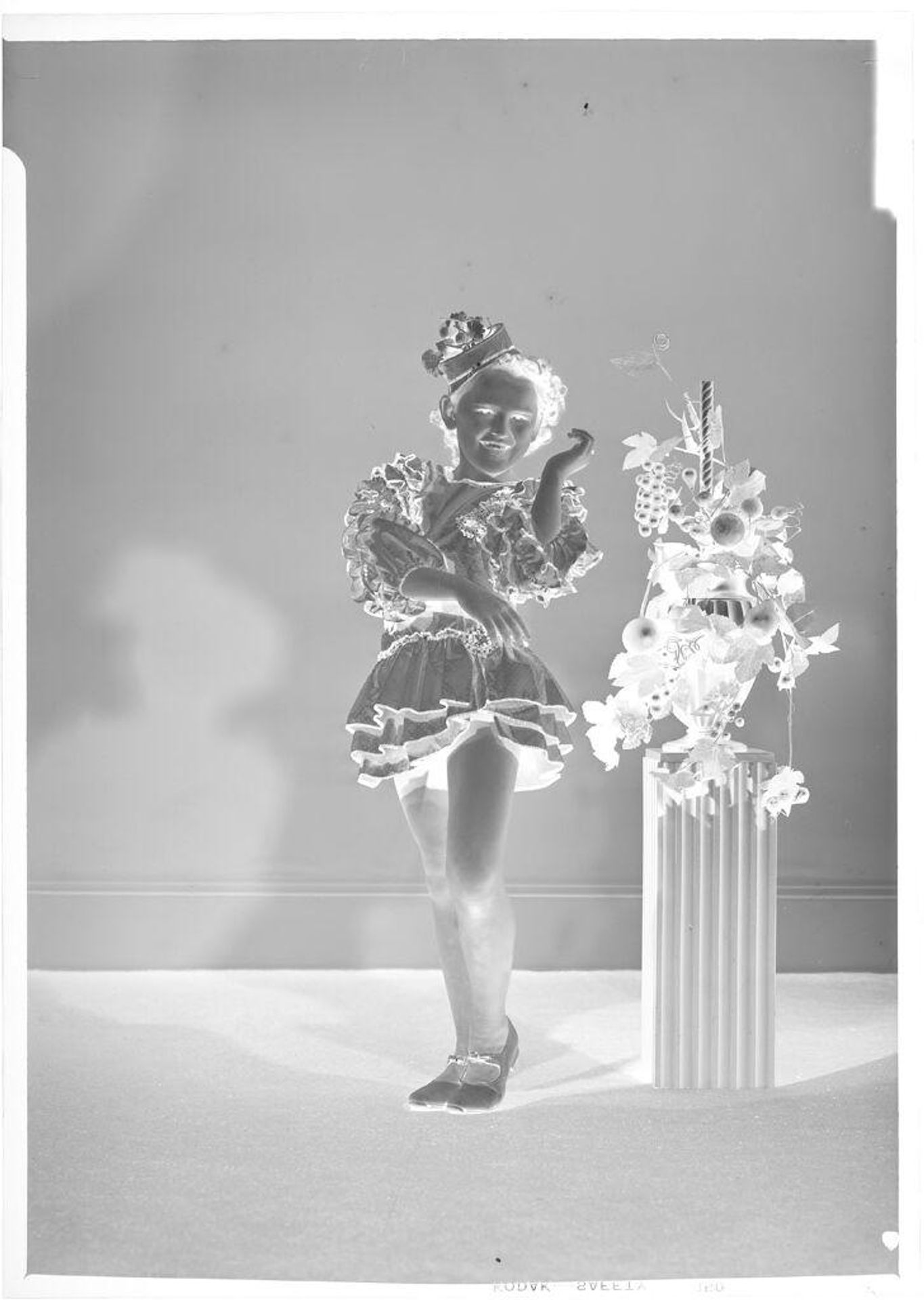 Untitled (girl posing in dance costume with tap shoes, next to basket of flowers atop column) by John Deusing