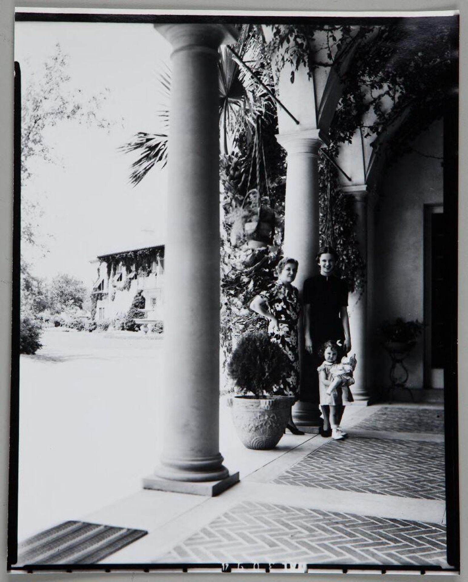 Untitled (two women with young girl holding doll standing on veranda by column) by Paul Gittings