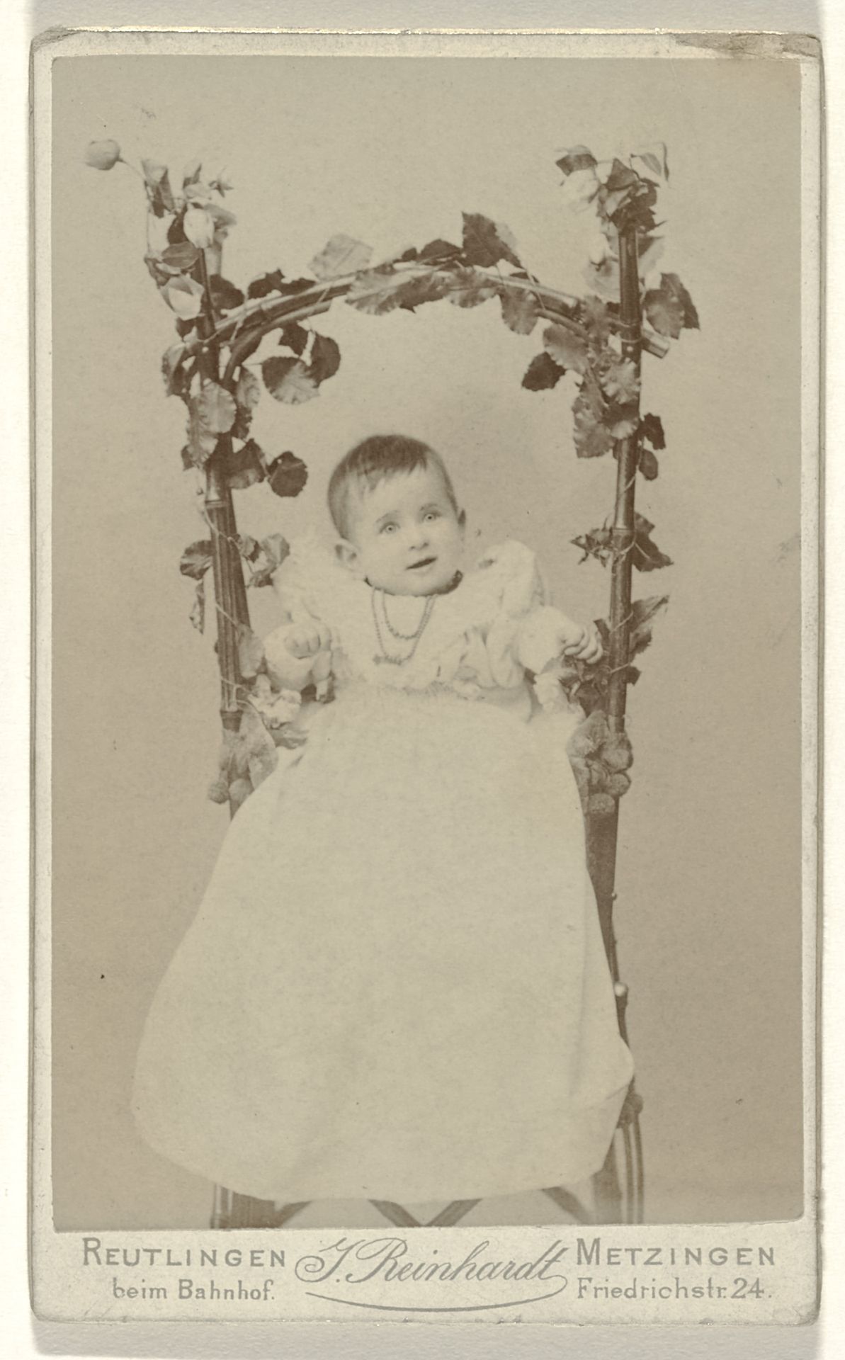 Jong kind, vermoedelijk Else Wachenheimer-Moos, in kinderjurk met kort haar en met een ketting om haar nek, in een kinderstoel met daarop bevestigd bladeren by J. Reinhardt