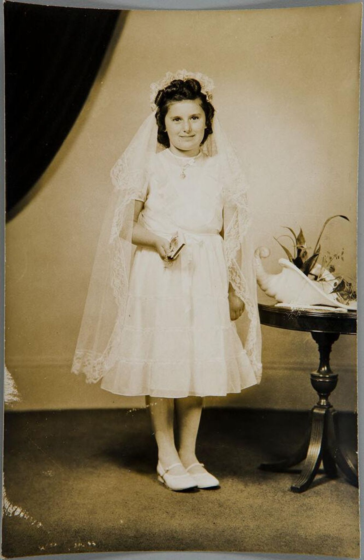 Untitled (girl standing in white, wearing veil, holding bible and rosary, one hand by side) by John Deusing