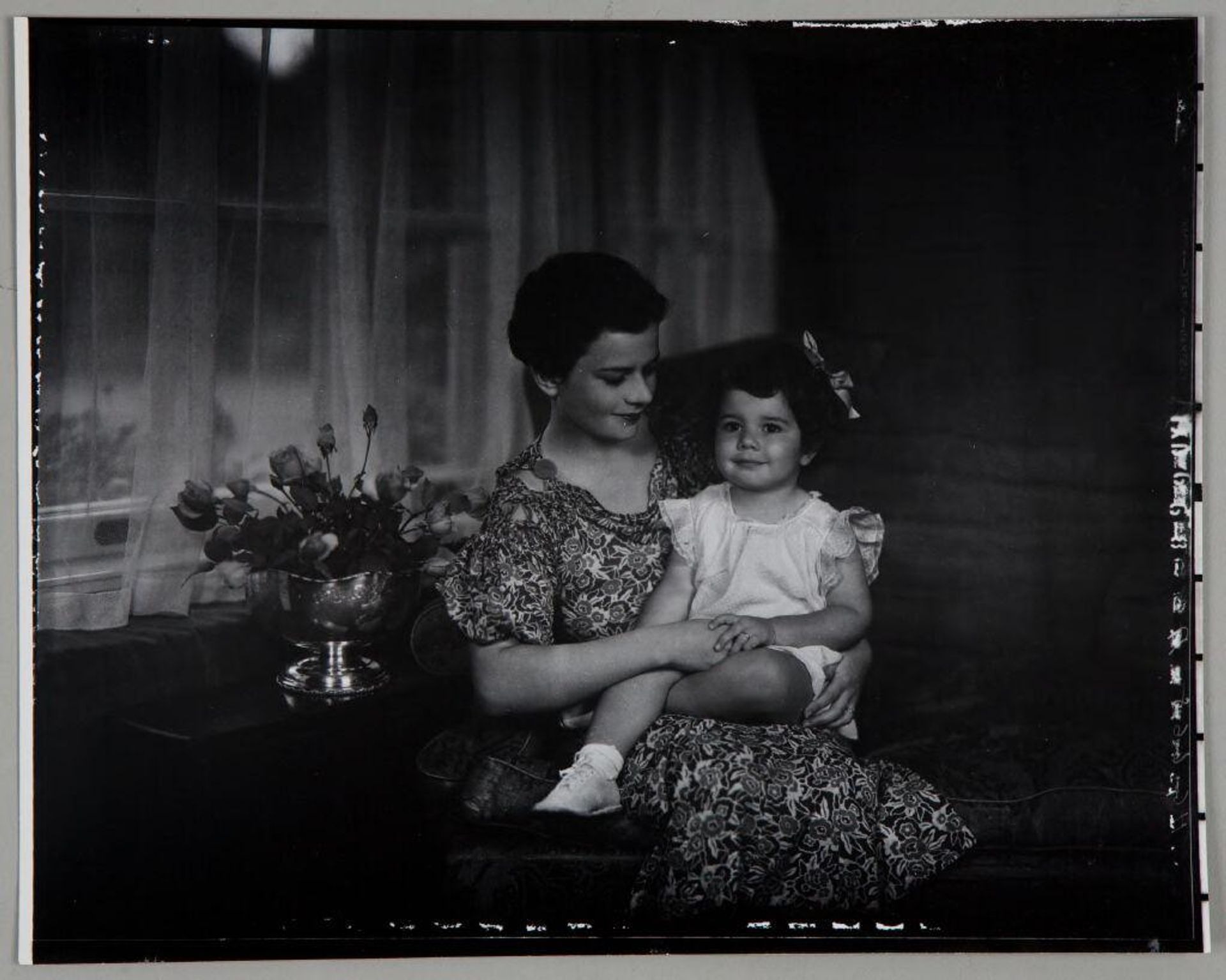Untitled (woman in floral dress with little girl on lap, holding hands, silver bowl of roses to left) by Paul Gittings