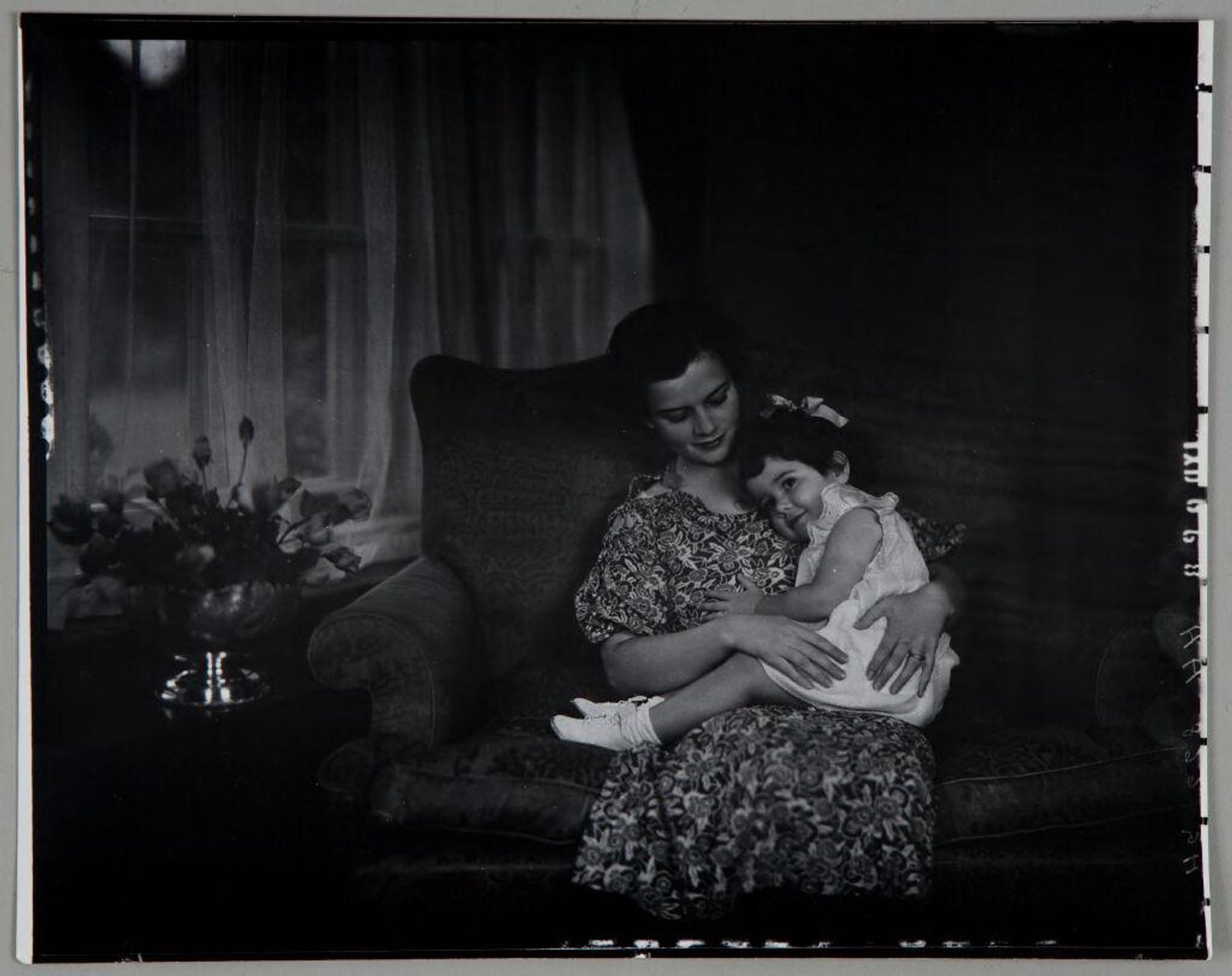 Untitled (woman in floral dress seated on sofa, little girl rests head on her shoulder) by Paul Gittings