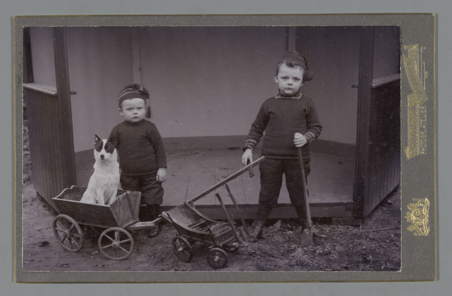 Portret van twee jongens met gebreide mutsen op bij speelgoedkarren en een hond by C. Sanders Szn