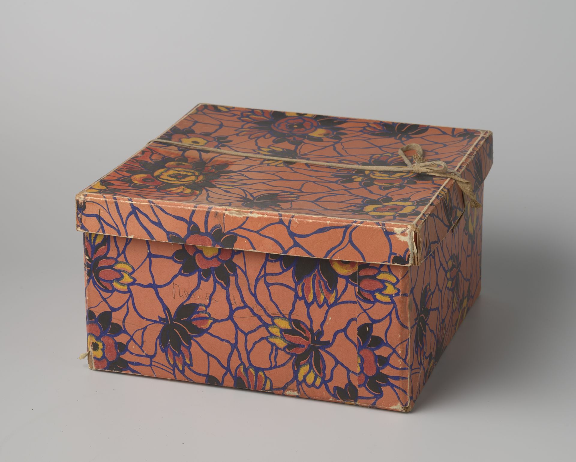 Vierkante hoedendoos van karton, beplakt met art deco papier met een oranje fond waarop een grillig patroon in blauw en bloemen in zwart, geel en rood by Maison Marcel