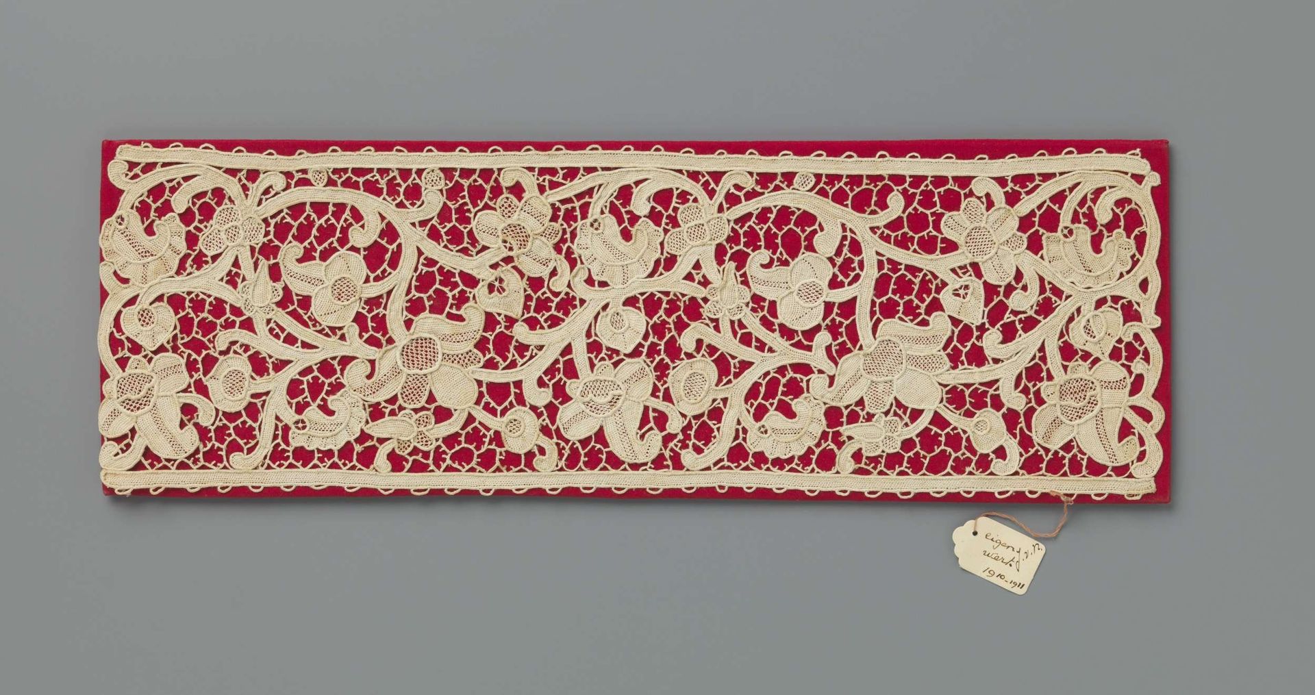 Strook naaldkant op een met rood textiel bekleed stuk karton by Johanna E. van Nierop
