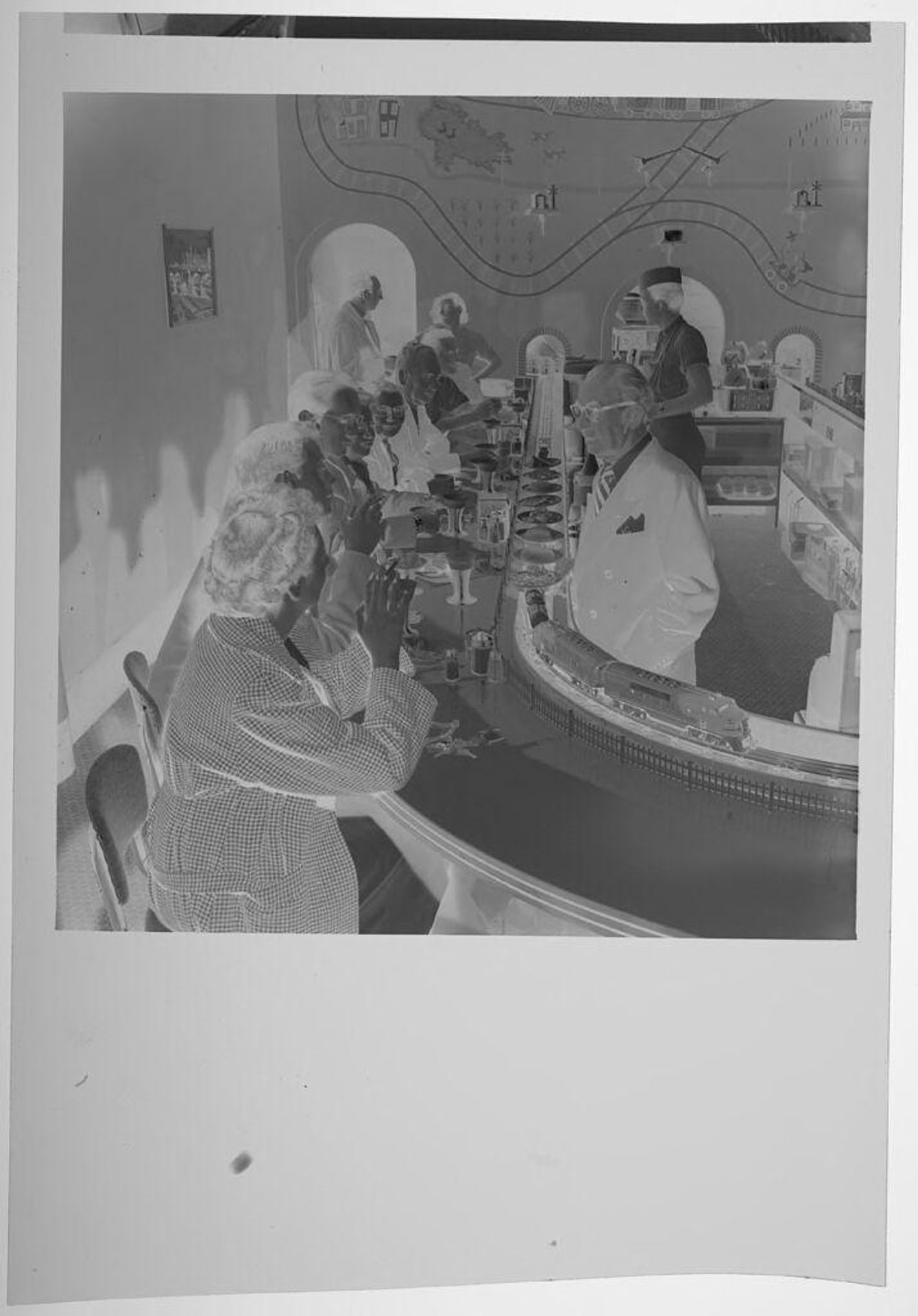 Untitled (people sitting at restaurant counter with circular model train) by Jack Gould