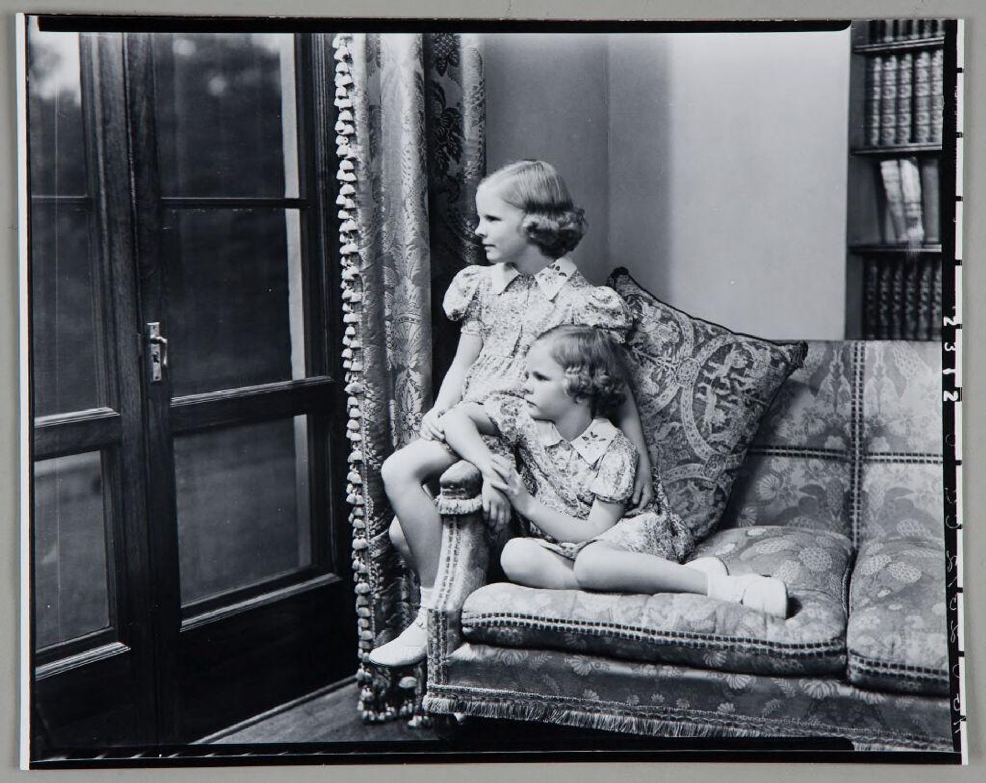 Untitled (two girls in matching floral dresses seated at edge of sofa looking out glass door) by Paul Gittings