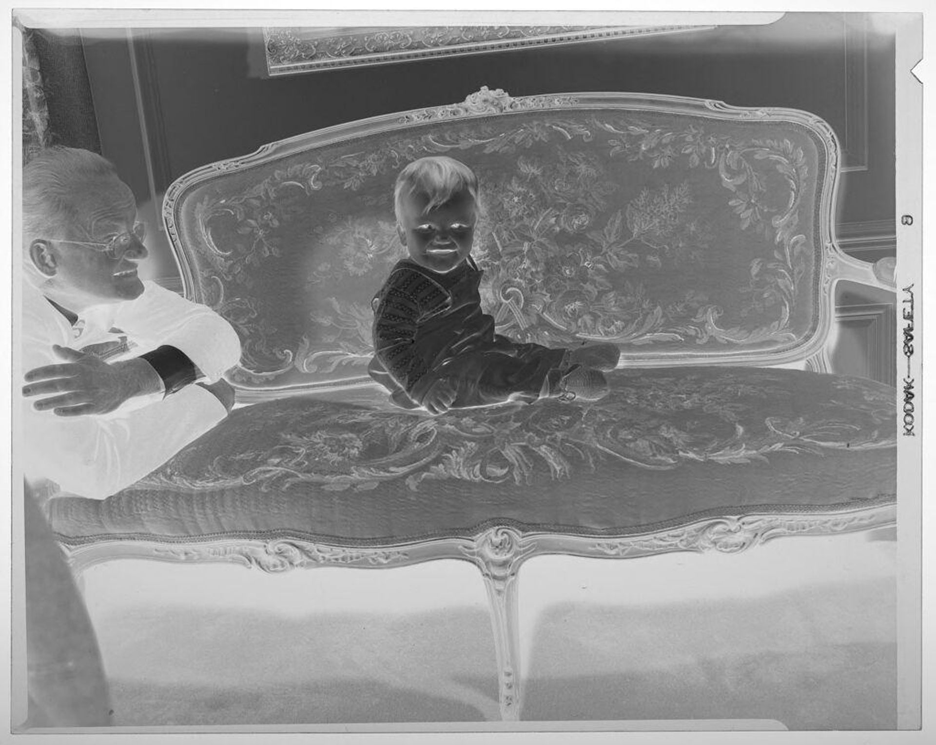 Untitled (baby posed sitting on ornate couch as grandfather looks on) by Martin Schweig