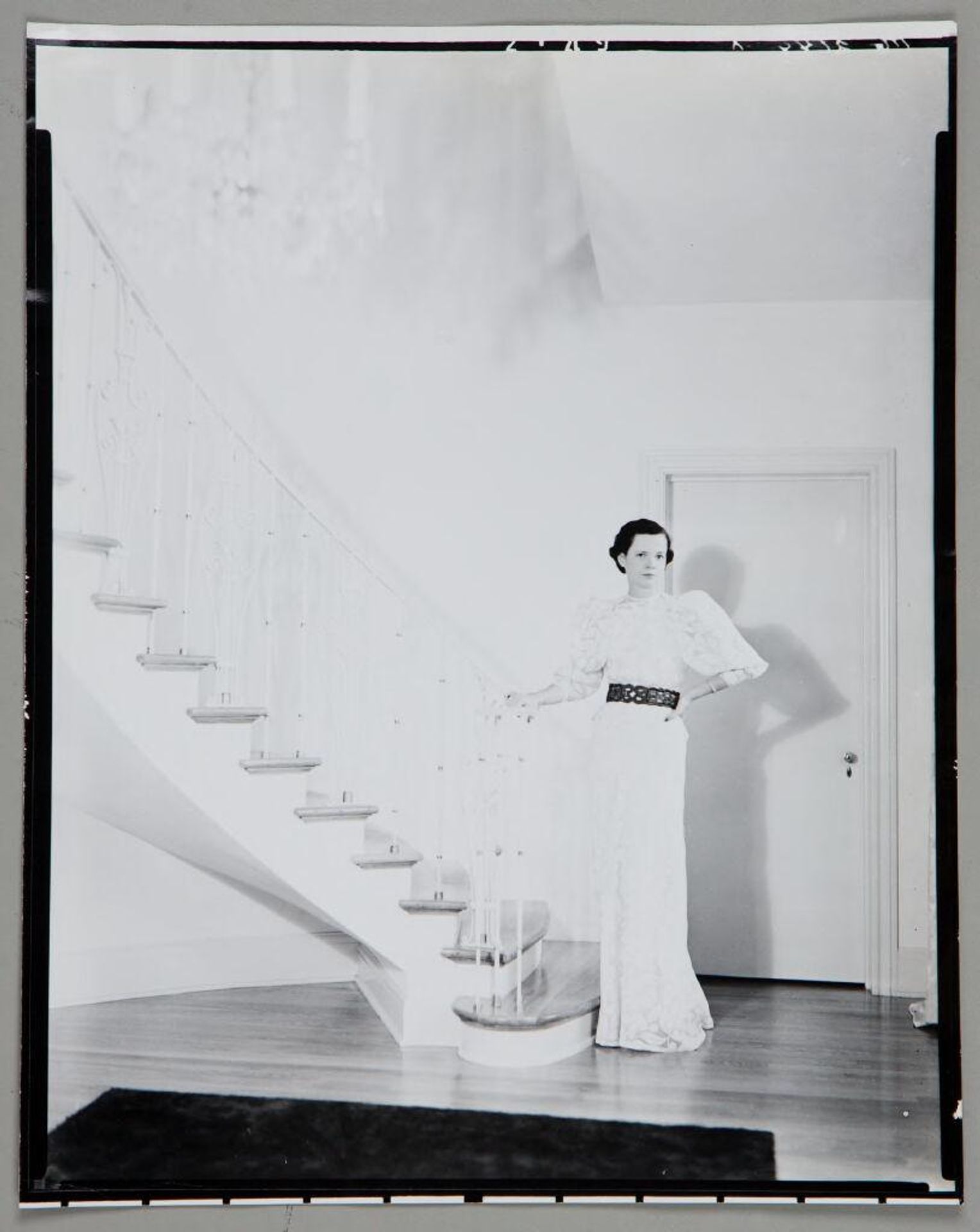 Untitled (young woman in long white dress with linked belt standing at base of curved staircase) by Paul Gittings