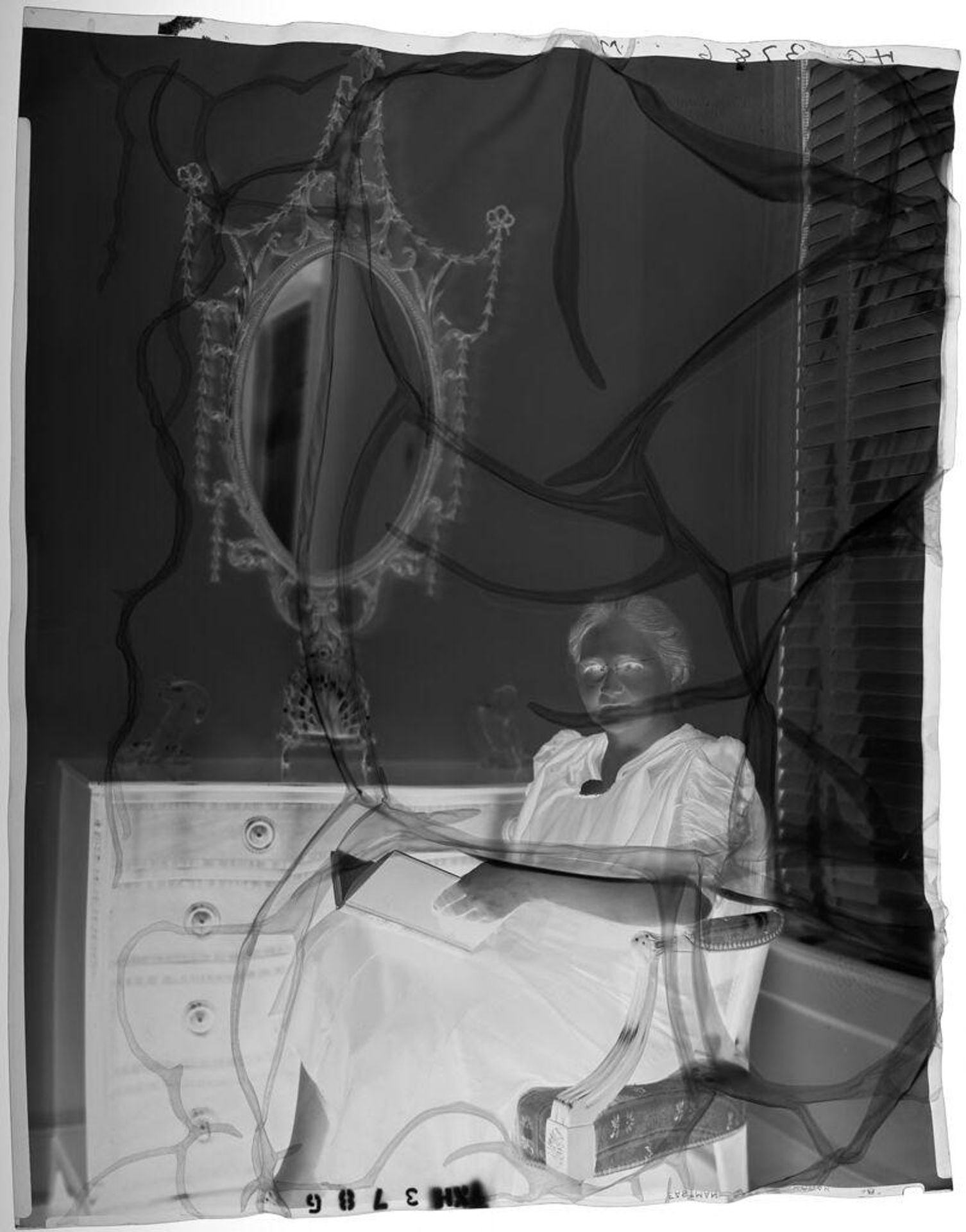 Untitled (older woman seated on chair in front of dresser and mirror, holding book) by Paul Gittings