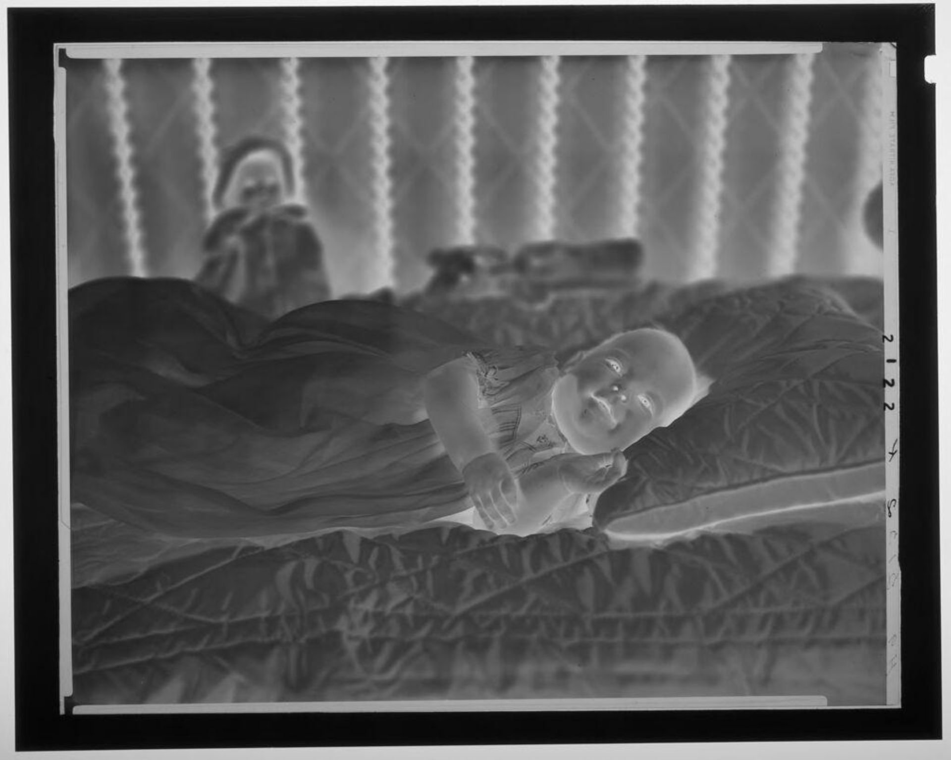 Untitled (baby in long dress lying on quilted pillow and cover) by Paul Gittings