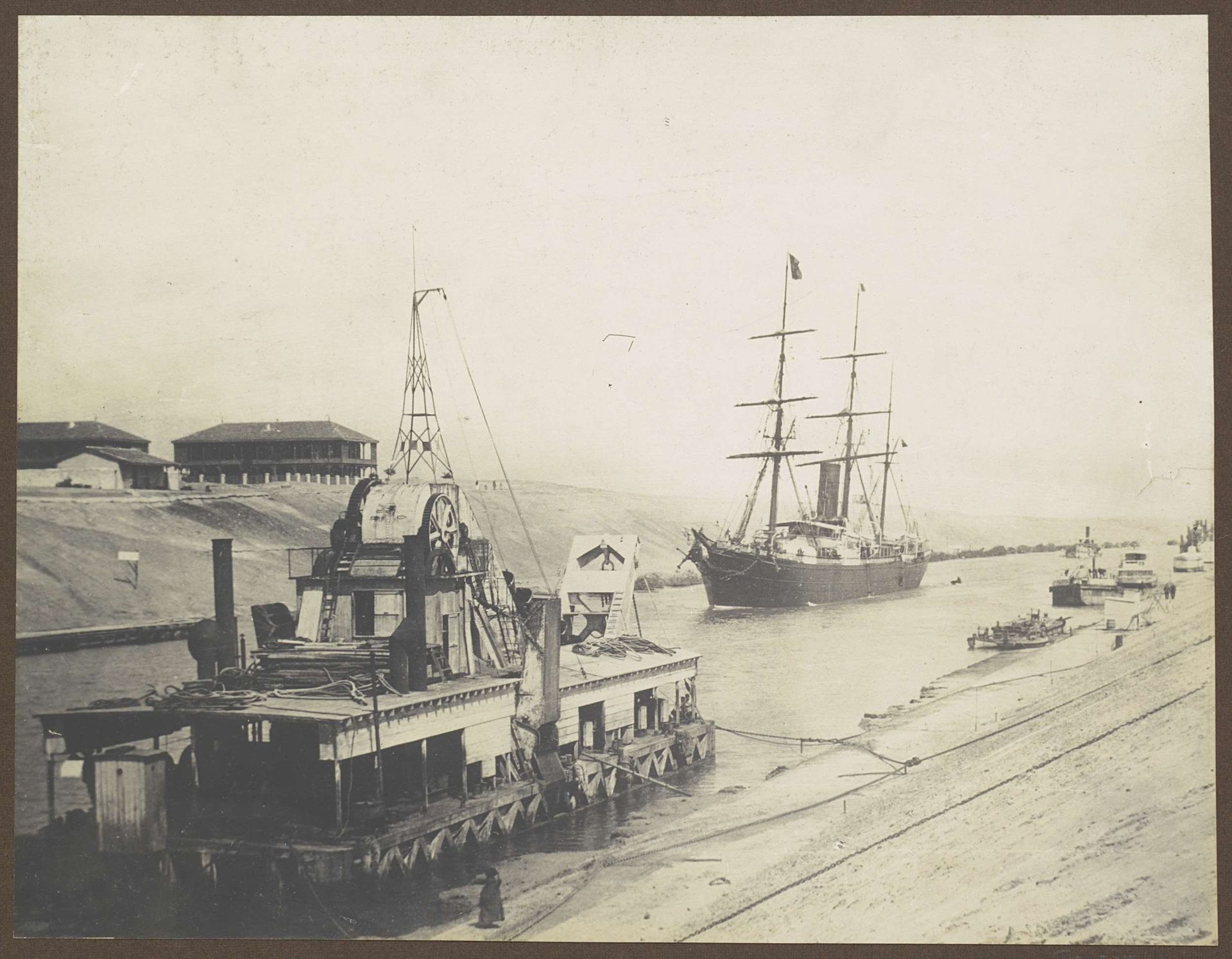 Schip vaart door het Suezkanaal bij een baggerschip en huizen by C. & G. Zangaki