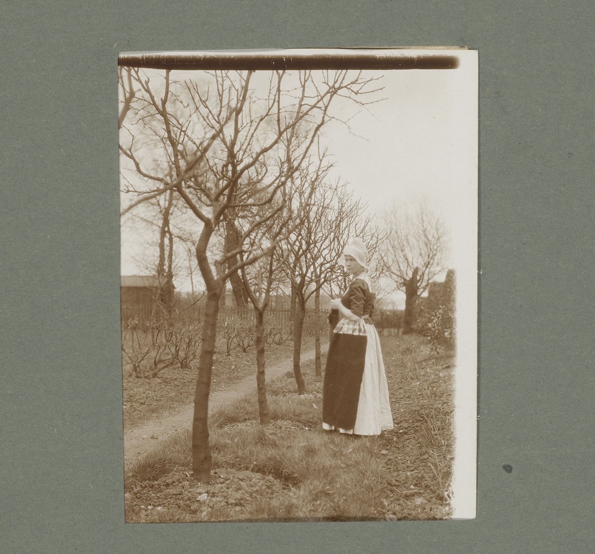 Vrouw in klederdracht met breiwerk, staande tussen kale bomen by G. Hidderley