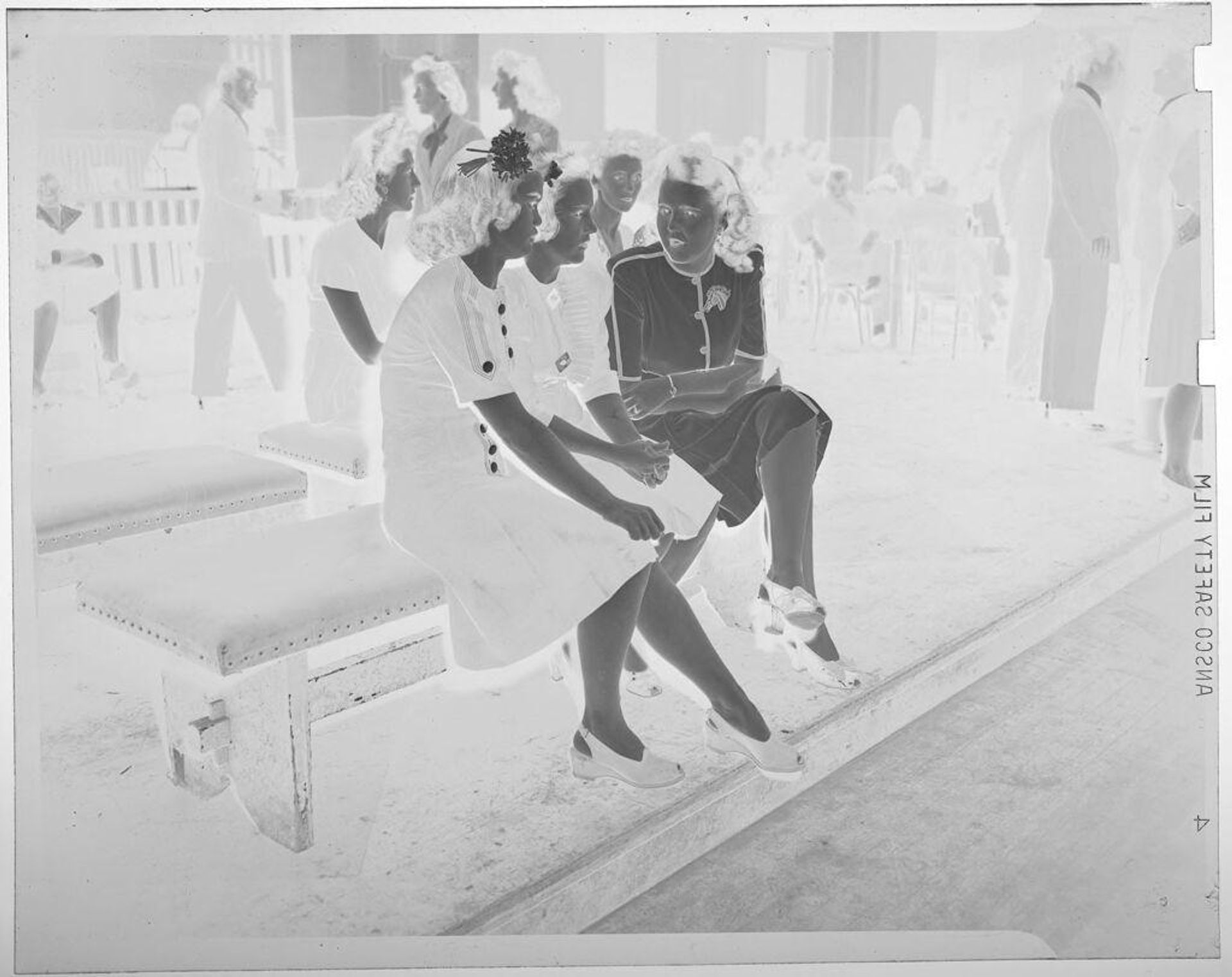 Untitled (three young women sitting on a bench, more benches and people in background) by Jack Gould