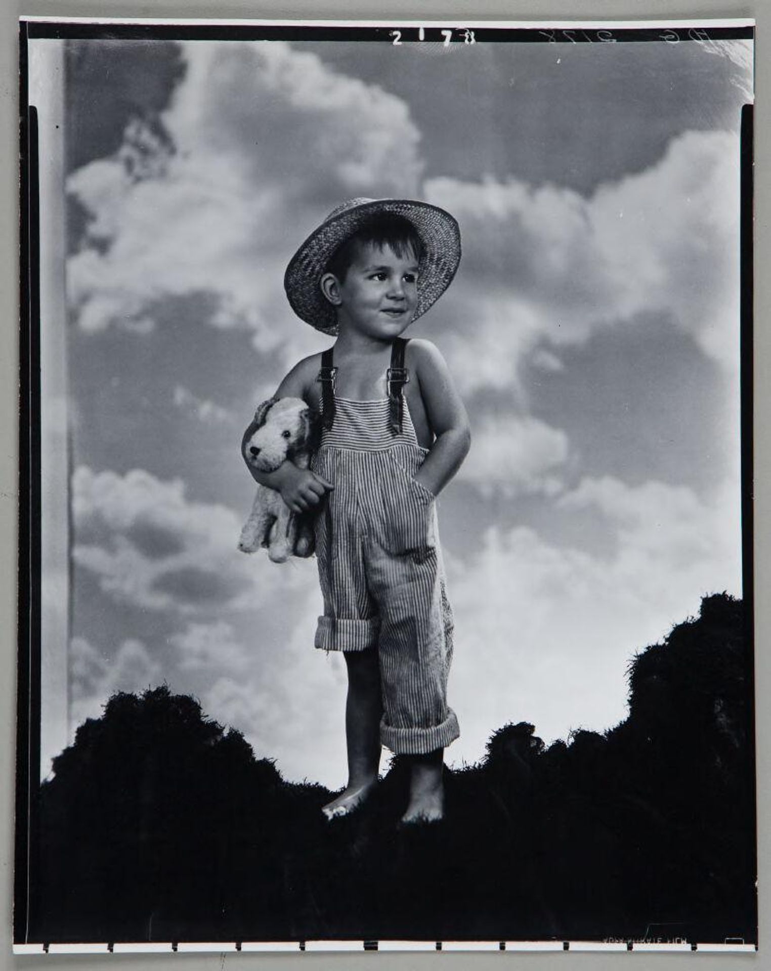Untitled (little boy in straw hat and striped overalls with stuffed dog under his arm) by Paul Gittings
