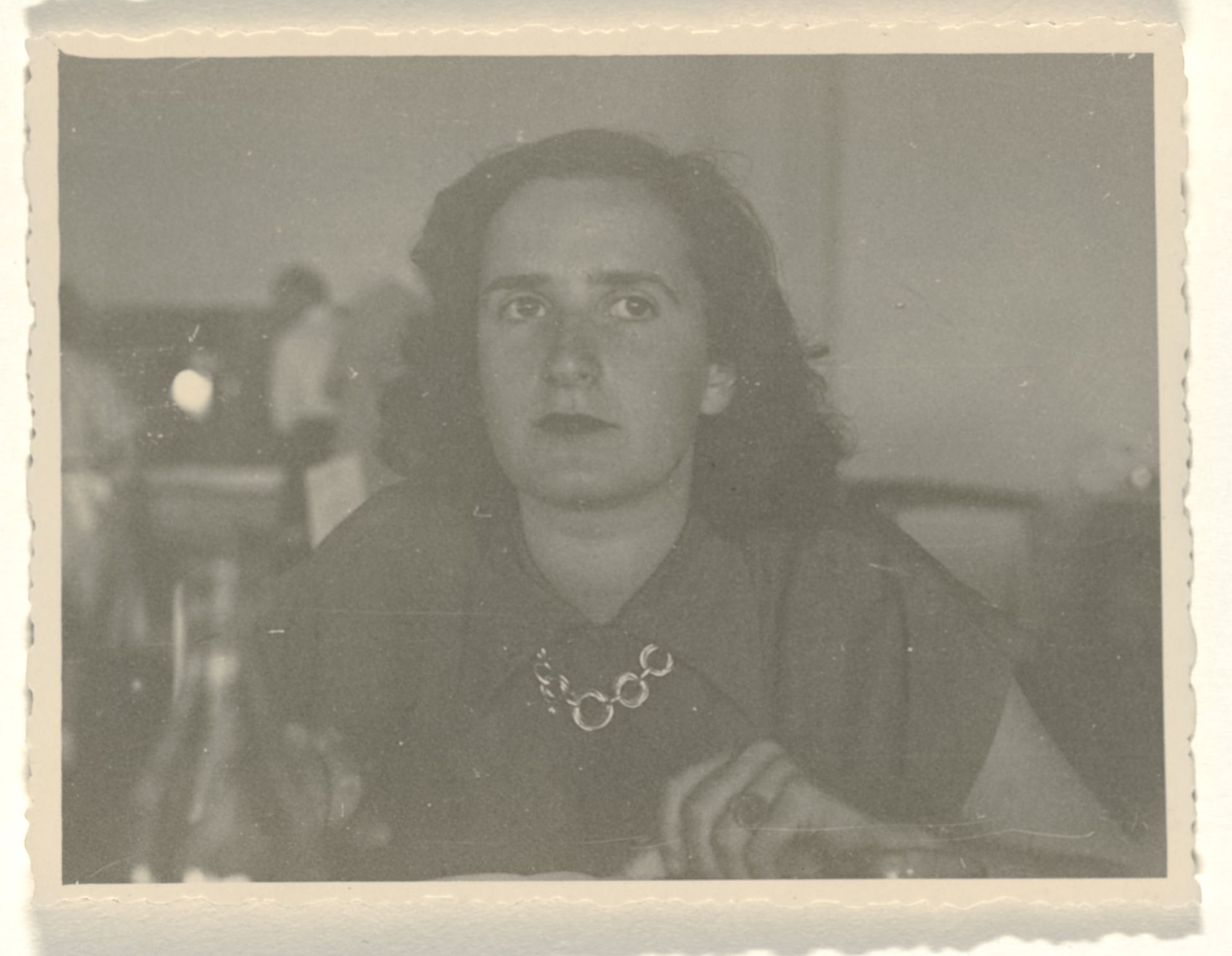 Isabel Wachenheimer zittend in zaal in hemd met korte mouwen met schakkelketting om haar hals, met onbekende mensen op de achtergrond by Anonymous