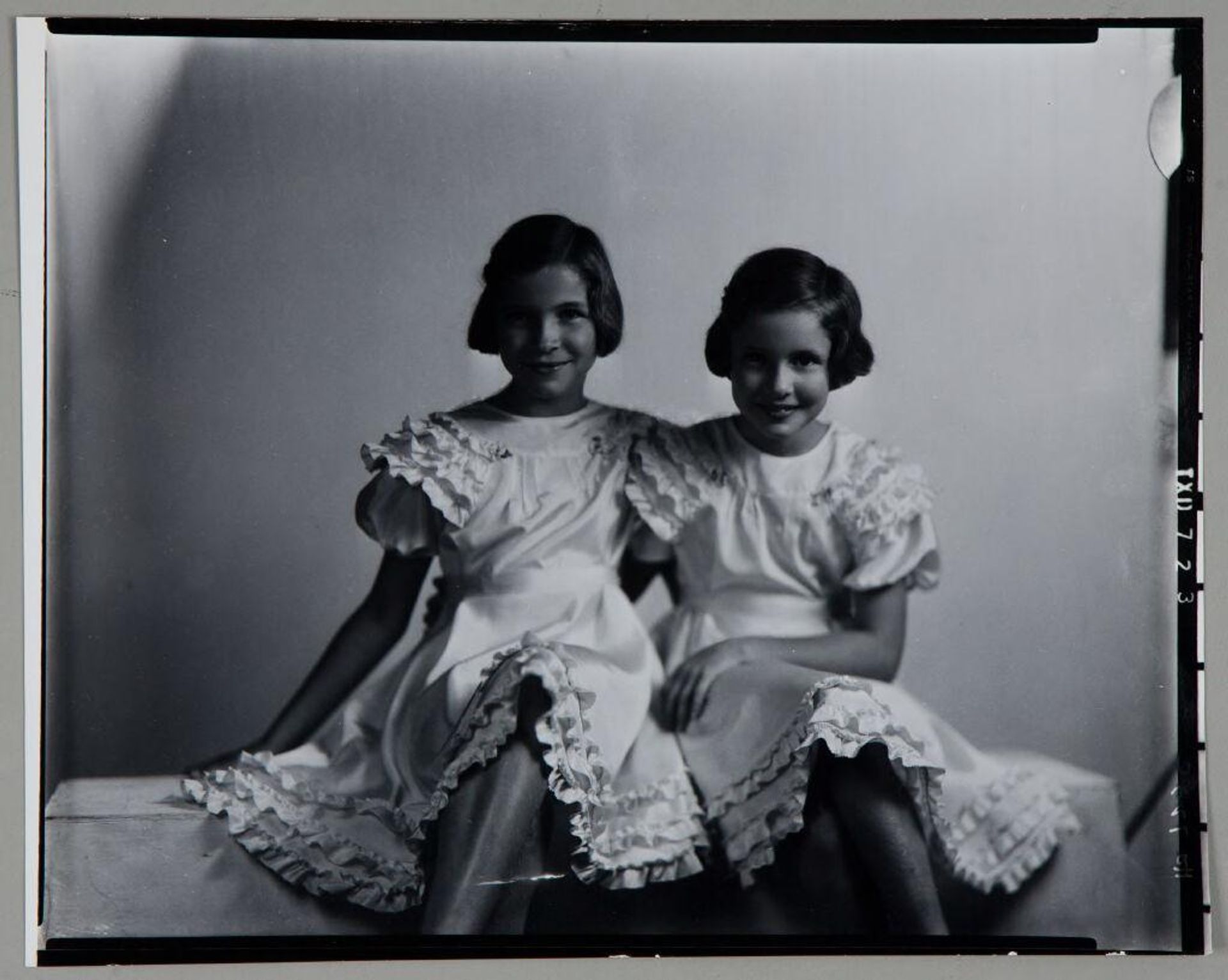Untitled (two girls in matching dreses seated in studio for portrait, legs crossed) by Paul Gittings