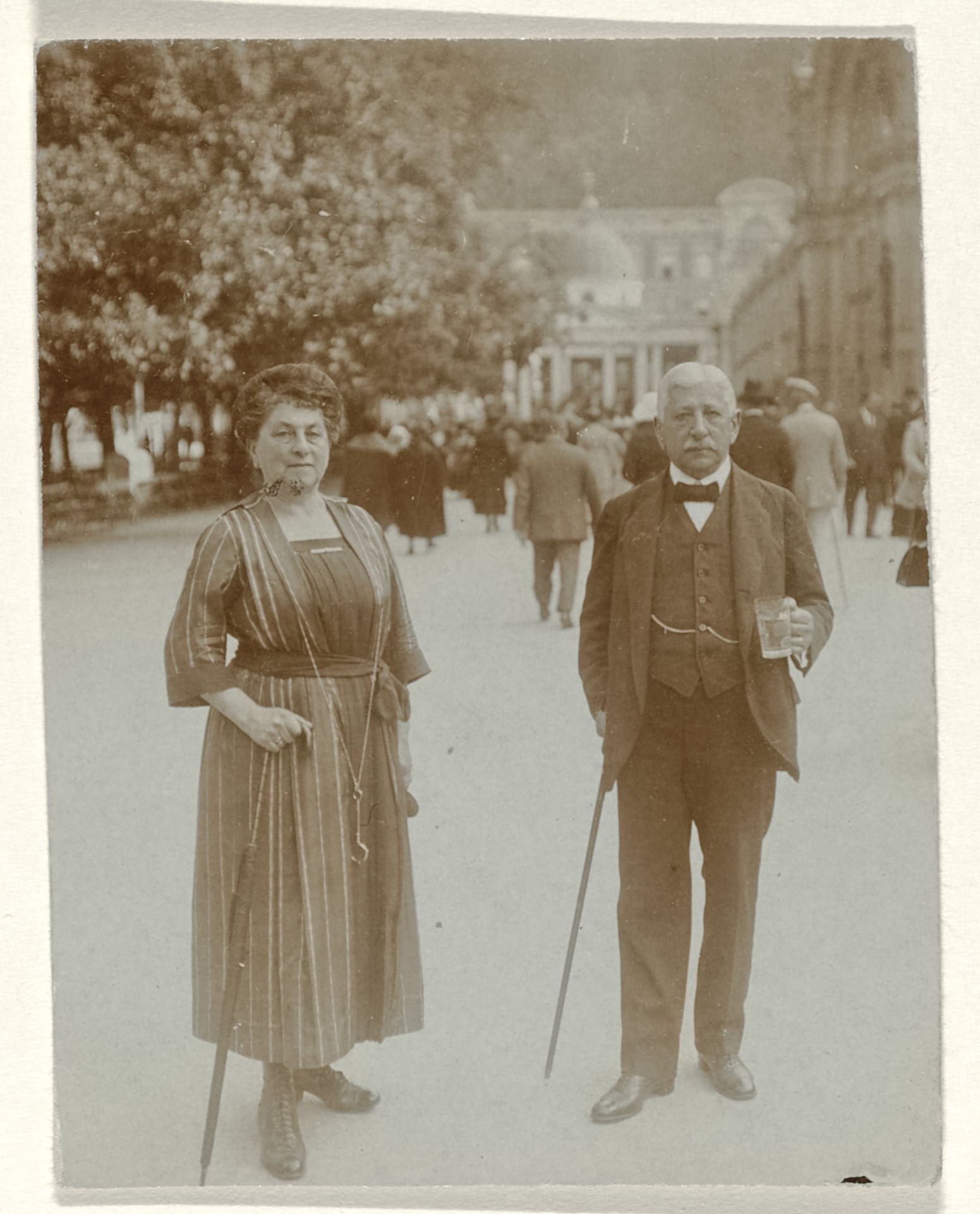Josefine Moos-Levi en haar echtgenoot Hermann Moos, beide met wandelstokken in hun handen en Hermann met een glas in zijn hand, staand op een straat met mensen, gebouwen en bomen by Anonymous