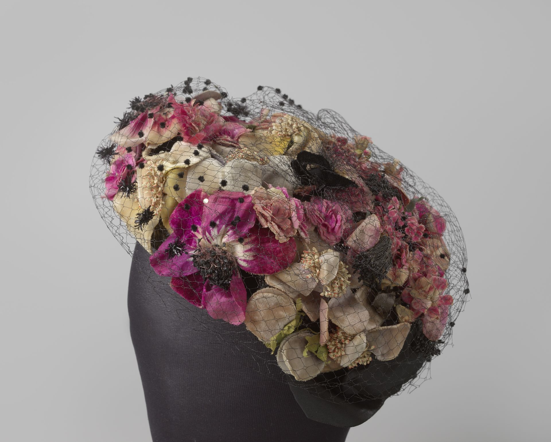 Hoed met zwarte strik in pillbox-model van zwart stro, geheel belegd met meerkleurige bloemen onder een afhangende zwarte voile met noppen en strasstenen by Wiener Mode