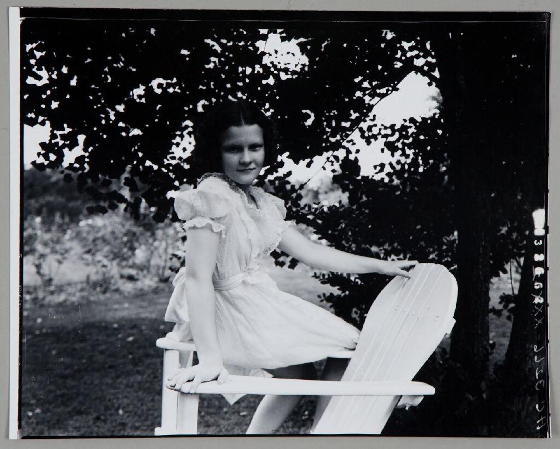 Untitled (girl in white dress seated on arm of wooden chair under tree) by Paul Gittings