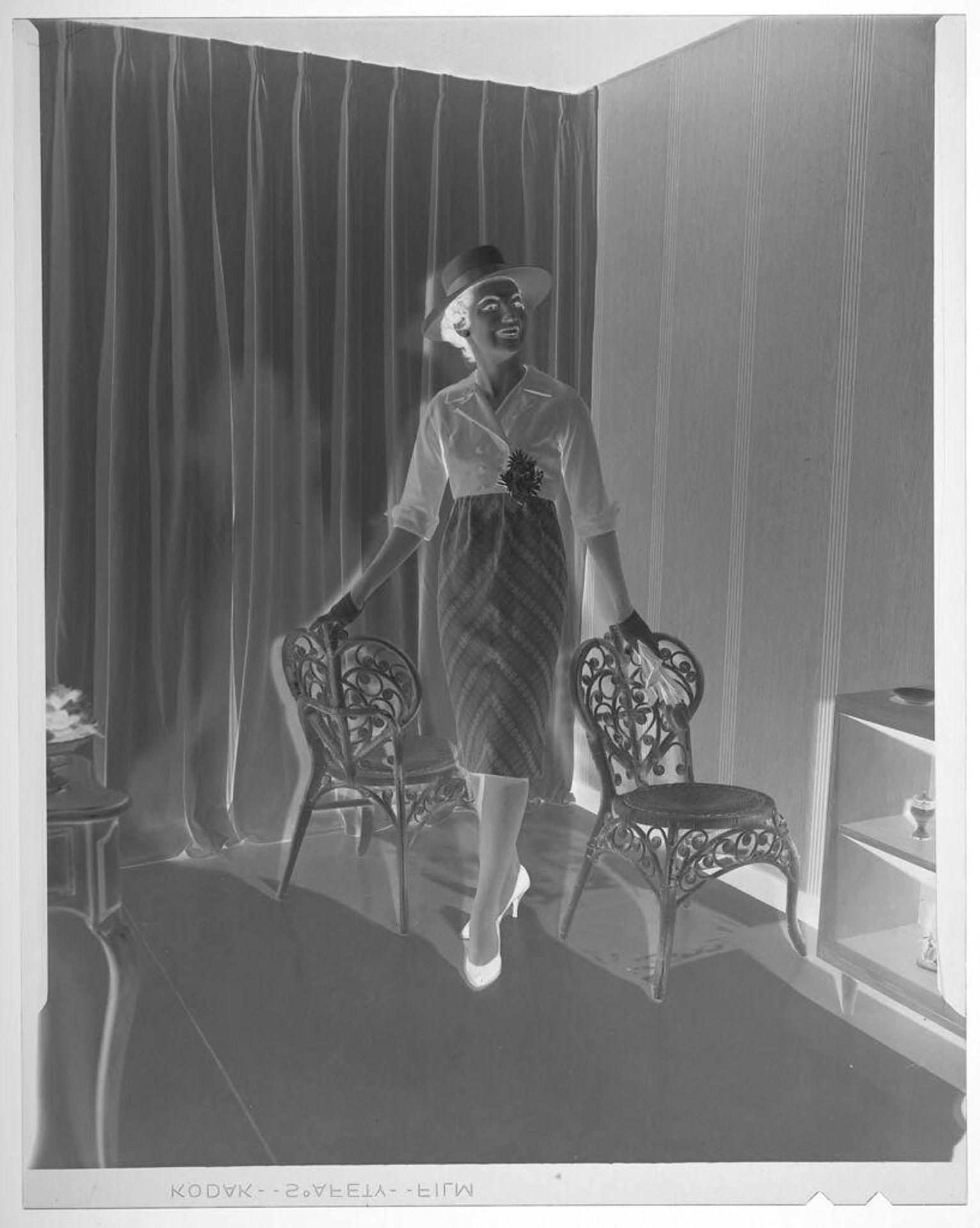 Untitled (fashion portrait of woman in hat and day clothes standing between two chairs) by Martin Schweig
