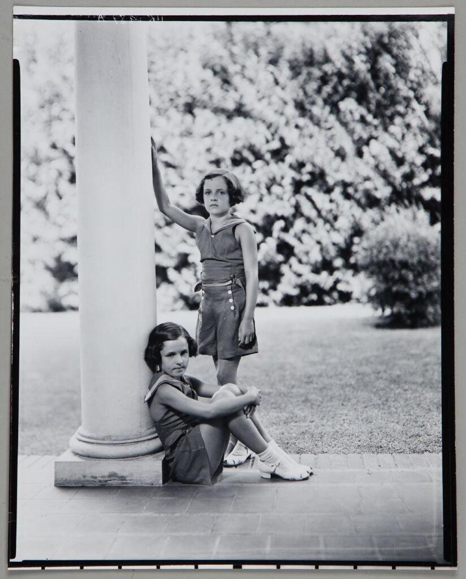 Untitled (twin girls in matching outfits on porch, standing and sitting against column) by Paul Gittings