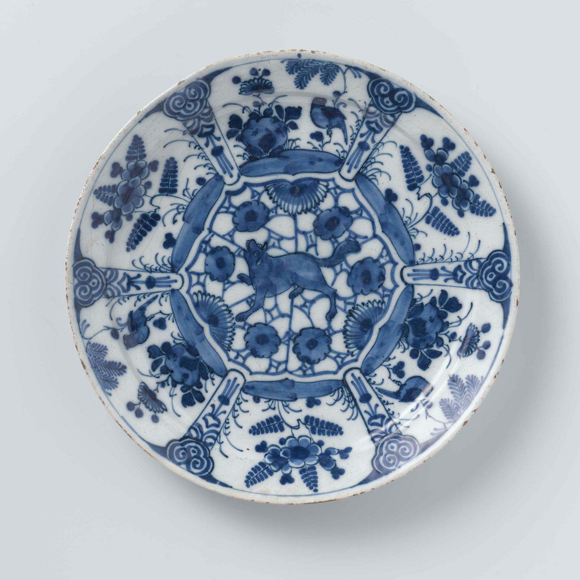 Bord van blauw en wit Delfts aardewerk, versierd met planten en vogels en een grommende wolf of vos in het midden by De Drie Posteleyne Astonne