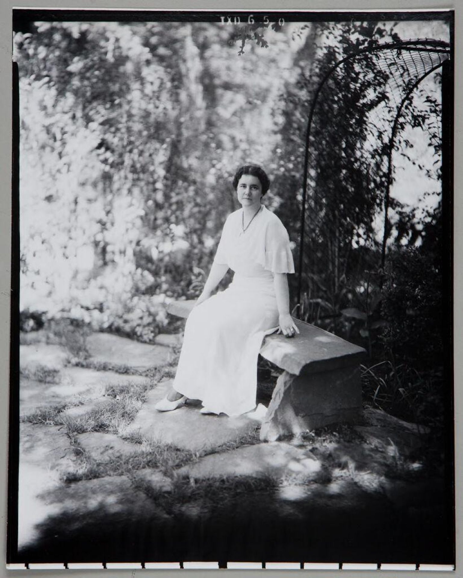 Untitled (woman in long white dress seated on stone bench in front of trellis) by Paul Gittings