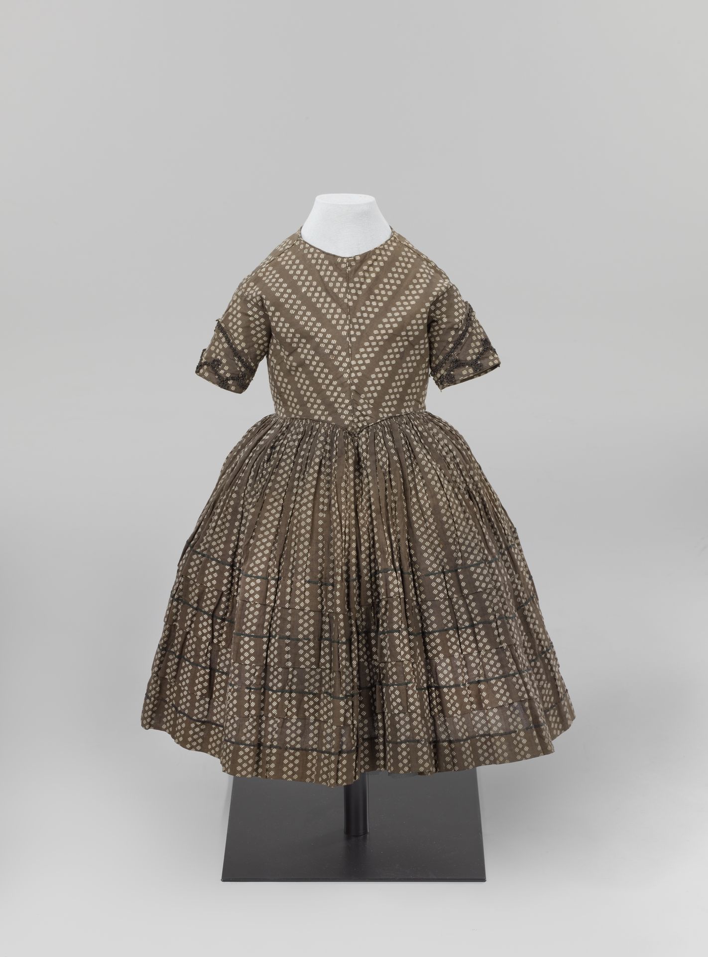 Japon van katoenen batist, bedrukt met een bruin fond waarin een verticaal gestreept strooipatroon is uitgespaard. Ronde hals, puntig lijf met balein middenvoor, gerimpelde wijde rok met drie oprijgen. Versierd met bruin passement en band. Lijf gevoerd met katoen. by Anonymous