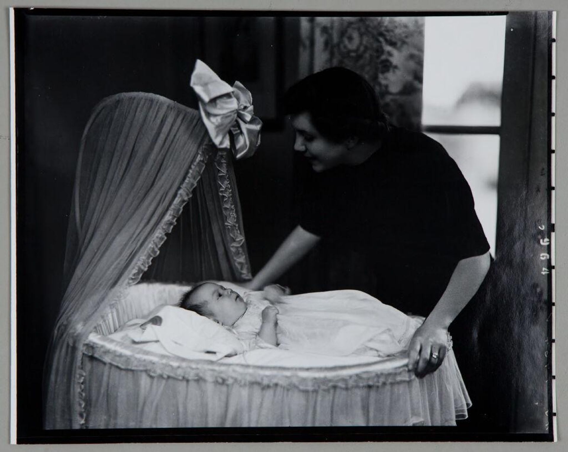 Untitled (woman leaning over baby in bassinet with large bow at top) by Paul Gittings