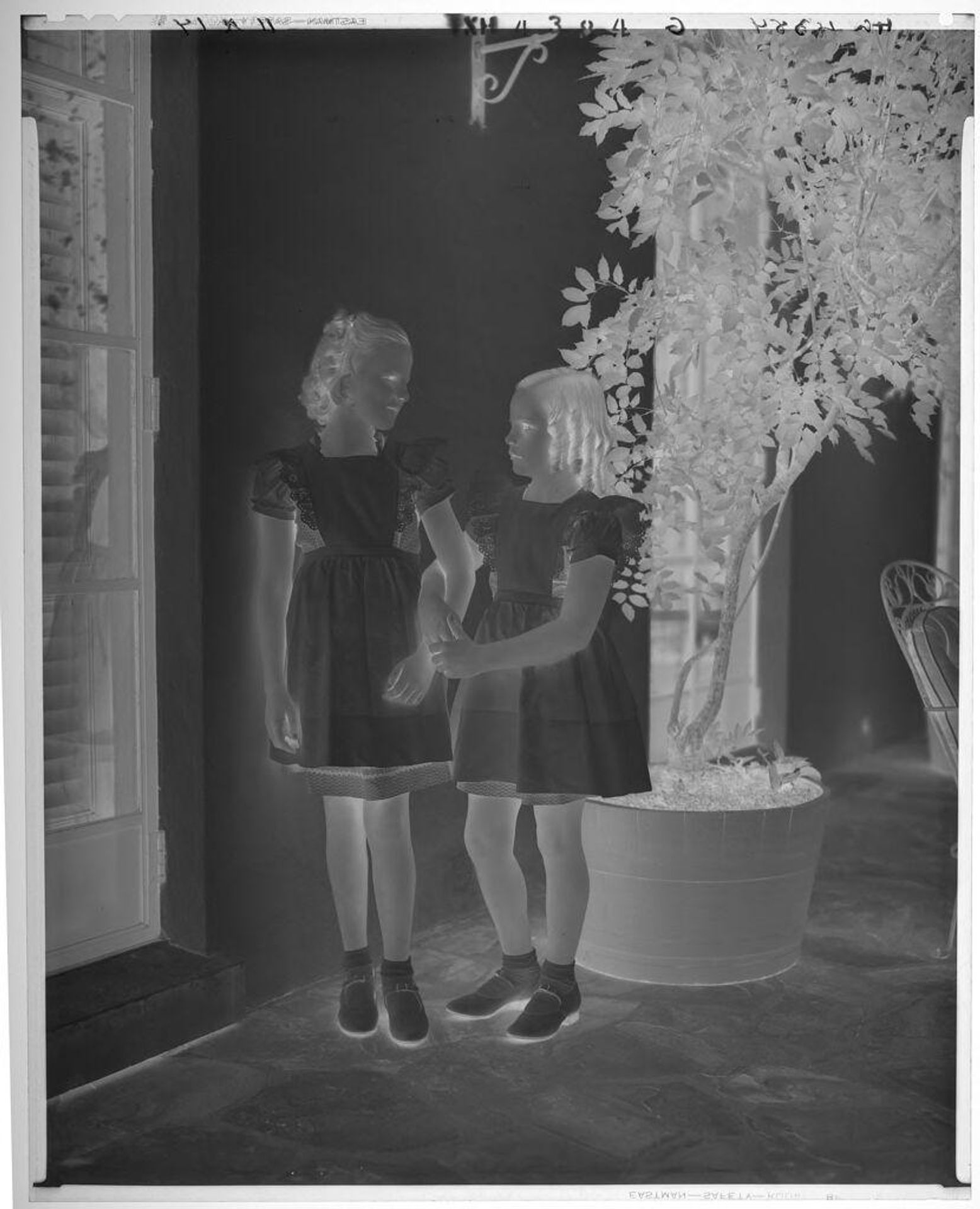 Untitled (two girls in matching dresses, arms interlocked, standing between vine and door on porch) by Paul Gittings