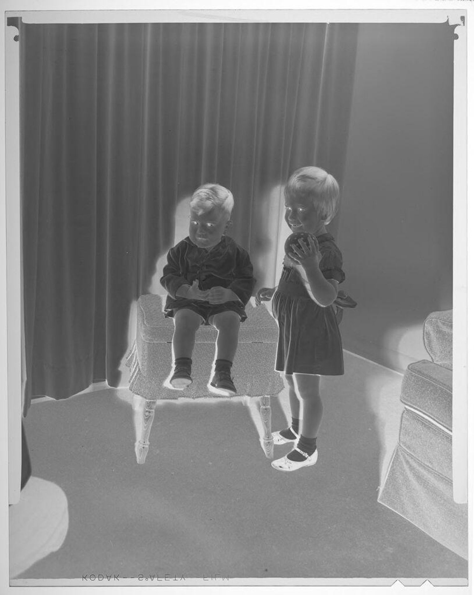 Untitled (young girl holding ball and young boy sitting on ottoman posing by curtained window) by Martin Schweig